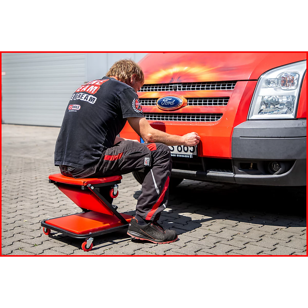 Ein Mann sitzt auf einem roten Rollhocker vor einem roten Ford-Transporter und arbeitet.